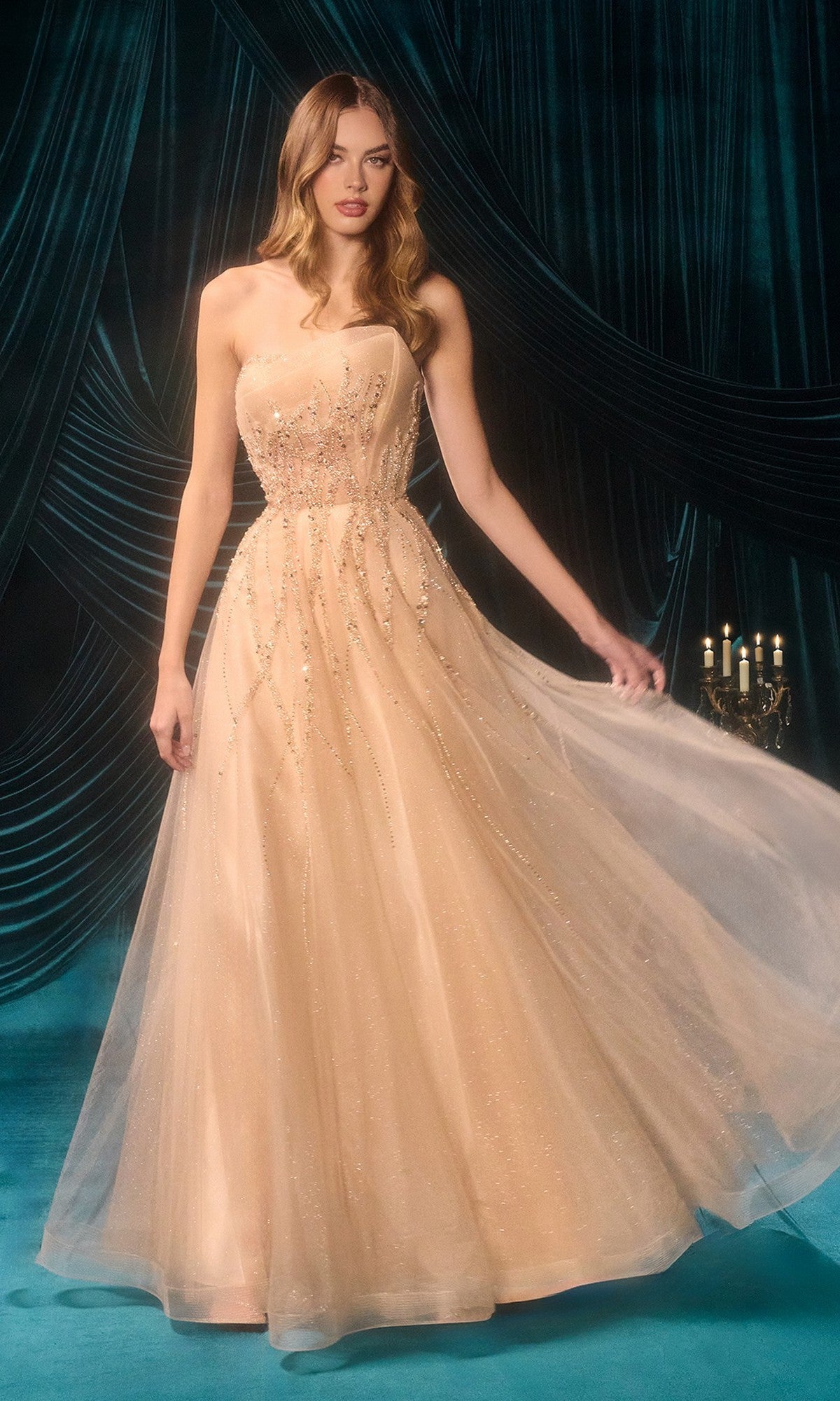 A woman wearing the champagne Long Prom Dress: Andrea and Leo A1491, a strapless floor-length gown with sparkling embellishments, stands in front of draped dark teal curtains.